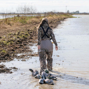 Person wearing camouflage waders walking through shallow water with duck decoys