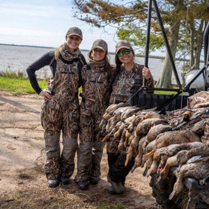 Three women in camouflage hunting gear standing by a truck bed filled with harvested ducks near a lake