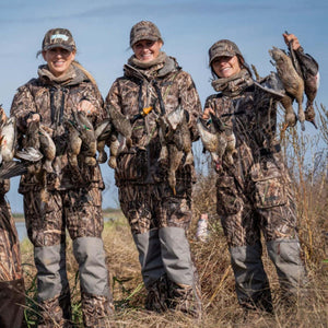 Three women wearing DSG Outerwear camouflage hunting gear holding ducks in a field