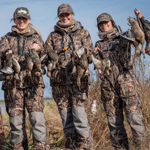 Three women wearing DSG Outerwear camouflage hunting gear holding ducks in a field