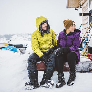 Man and woman wearing winter jackets and Baffin mittens sitting on a wooden porch in the snow