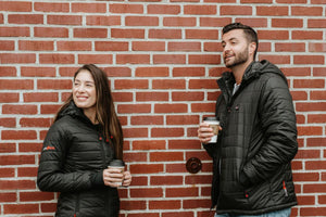 Man and woman wearing black ActionHeat puffer jackets standing against a brick wall holding coffee cups