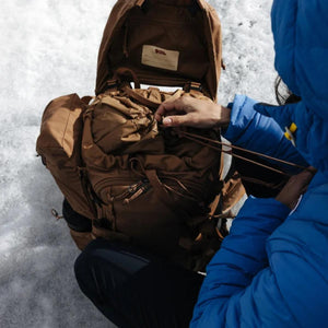 Person in blue jacket adjusting the drawstring on a brown FjallRaven backpack in the snow