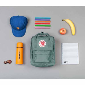 Fjallraven Kanken backpack with blue cap, Primus bottle, markers, notebook, apple, banana, and nuts on grey background
