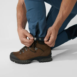Person adjusting the snap button on the hem of blue hiking trousers over brown leather hiking boots