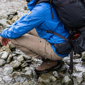 Person wearing Fjallraven blue jacket and beige trousers crouching on rocky stream bank with black backpack