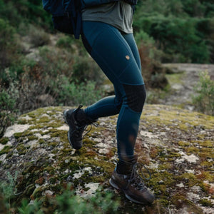 Person wearing blue Fjallraven hiking leggings and brown boots walking on a mossy rock