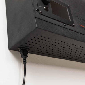 Black power cord plugged into the underside of a black electronic device with a perforated metal casing
