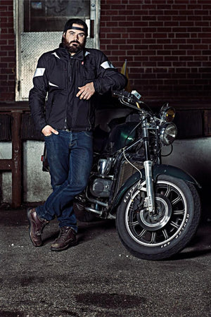 Man with beard wearing a black motorcycle jacket and cap leaning against a green motorcycle