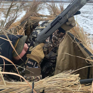 Hunter in camouflage gear wearing Glacier Glove waterproof gloves holding a shotgun in a grass blind