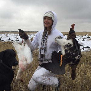 Woman wearing Glacier Glove gear holding two geese in a field with a black dog