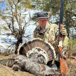 Hunter wearing Glacier Glove camouflage gear with a wild turkey in a wooded area