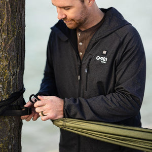 Man wearing a Gobi Heat black hooded jacket attaching a hammock to a tree