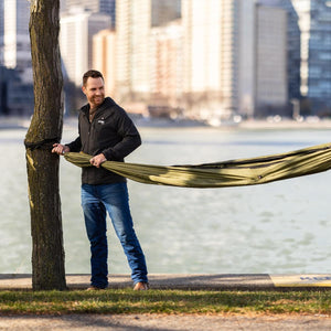 Man wearing a Gobi Heat jacket attaching a green hammock to a tree by a river