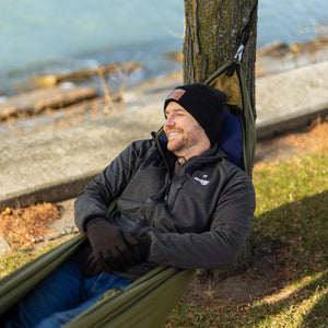 Man wearing Gobi Heat jacket and beanie resting in a hammock by a tree