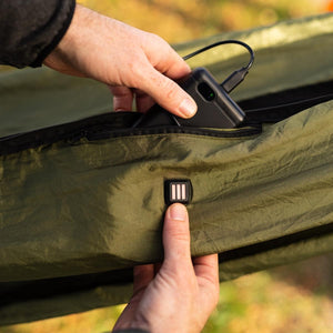 Hand connecting a power bank to the integrated heating control button on a green camping hammock