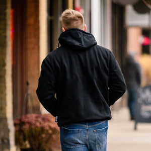 Person seen from behind wearing a black hooded sweatshirt and blue jeans walking on a sidewalk