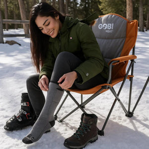 Woman wearing Gobi Heat jacket and heated socks sitting in a folding chair in the snow