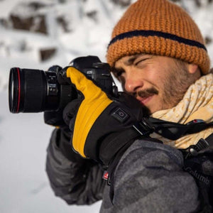 Person wearing Gobi Heat black and yellow gloves while holding a camera in a snowy environment