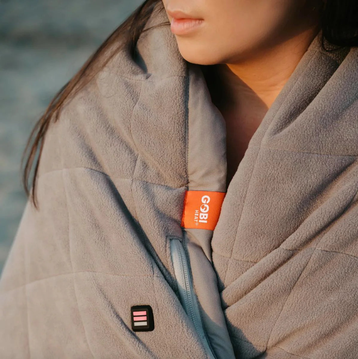Person wrapped in a grey Gobi Heat fleece blanket with a visible power button and brand tag
