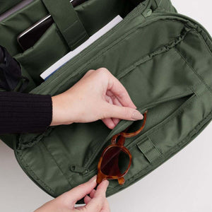 Hand placing tortoiseshell sunglasses into the interior zippered pocket of a green Got Bag backpack