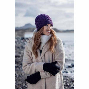 Woman wearing a purple cable knit beanie and black gloves with a beige coat by the coast