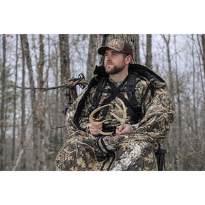Hunter in camouflage suit holding deer antlers while sitting in a tree stand with a compound bow