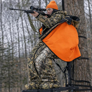 Hunter wearing Heater Body Suit camouflage gear and orange hat in a tree stand aiming a rifle