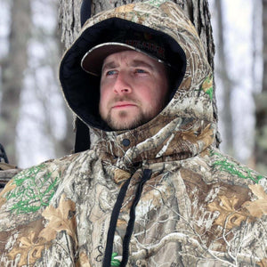 Man wearing a Heater Body Suit camouflage hunting parka with hood up in a snowy forest