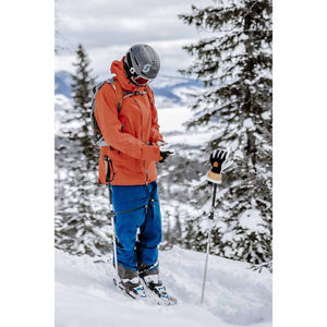 Skier wearing orange jacket and blue pants adjusting gloves in snowy mountain setting