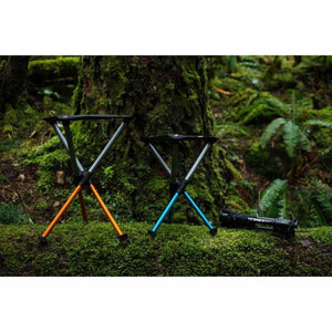 Two Hillsound portable stools in orange and blue standing on a mossy log with a folded stool nearby