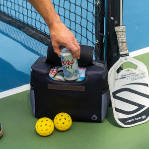 Hand reaching into a black Kanga Coolers bag on a pickleball court next to a paddle and two yellow balls