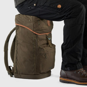 Person sitting on a dark olive FjallRaven backpack wearing black trousers and brown leather hiking boots