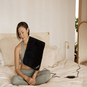 Person sitting on a bed using a black Mhigh electric heating pad on their shoulder