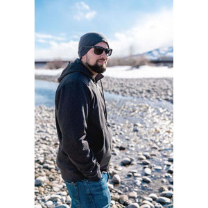 Man wearing a Mobile Warming charcoal grey hoodie and beanie standing by a rocky riverbank