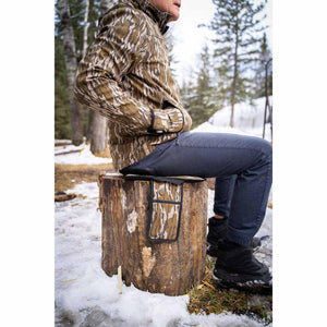 Person wearing Mobile Warming camouflage jacket sitting on a tree stump with a heated seat cushion