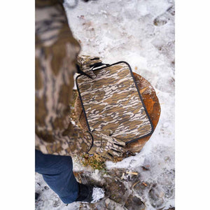 Person in camouflage gear holding a Mobile Warming camo seat pad on a tree stump in the snow