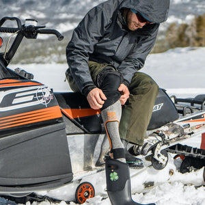 Man sitting on a snowmobile pulling on a Mobile Warming heated sock