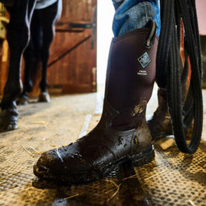 Muck brown rubber chore boot worn in a stable with horse legs visible in background