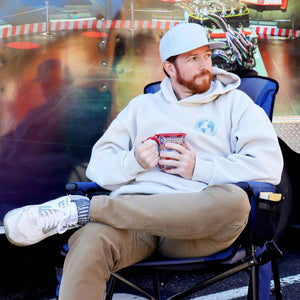 Man wearing ExtremeMIST hoodie and cap sitting in a folding chair holding a patterned mug