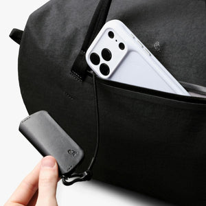 Bellroy black duffel bag with a white phone in the pocket and a hand holding a black leather key cover