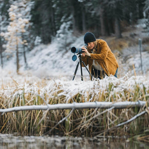 Person in brown jacket and beanie using a spotting scope on a tripod in a snowy forest