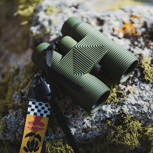Olive green NOCS Provisions binoculars with a patterned strap resting on a mossy rock
