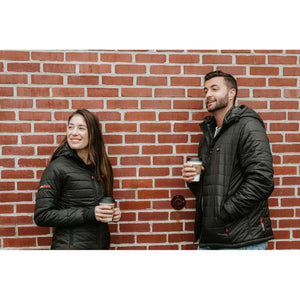 Man and woman wearing black ActionHeat hooded jackets standing against a brick wall holding coffee cups