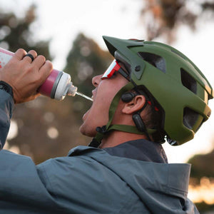Cyclist wearing an olive green helmet and Outdoor Tech bone conduction headphones drinking from a water bottle