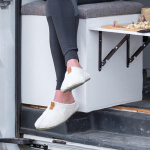 Person wearing white Acorn faux fur slippers while sitting on a van bench