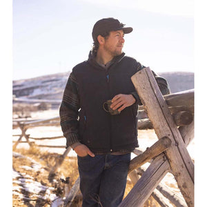 Man wearing a dark fleece vest and cap holding a mug while leaning against a wooden fence in a snowy field
