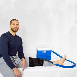 Man wearing a black knee compression wrap connected to a blue portable ice therapy cooler