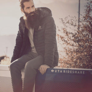 Man with beard wearing a black Ravean down jacket sitting on a UTA Rideshare bench
