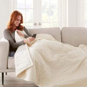 Woman sitting on a sofa using a smartphone while covered with a cream colored electric heated blanket